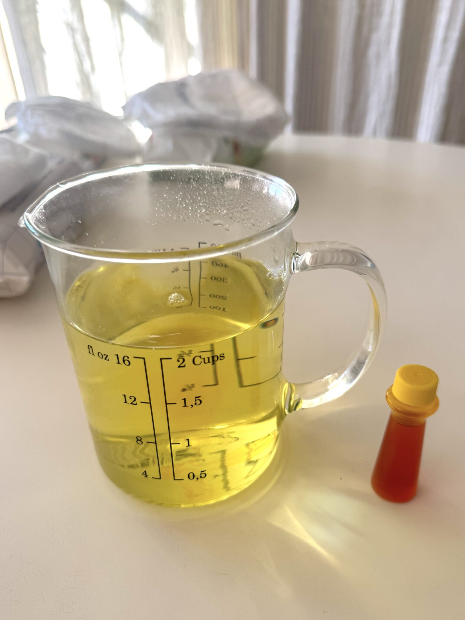 A glass measuring cup with yellow liquid sits on a white surface next to a small plastic bottle with a yellow cap.