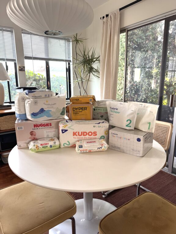 A round white table displays various packs of baby diapers and wipes from multiple brands in a sunlit room with large windows and a hanging lamp above.