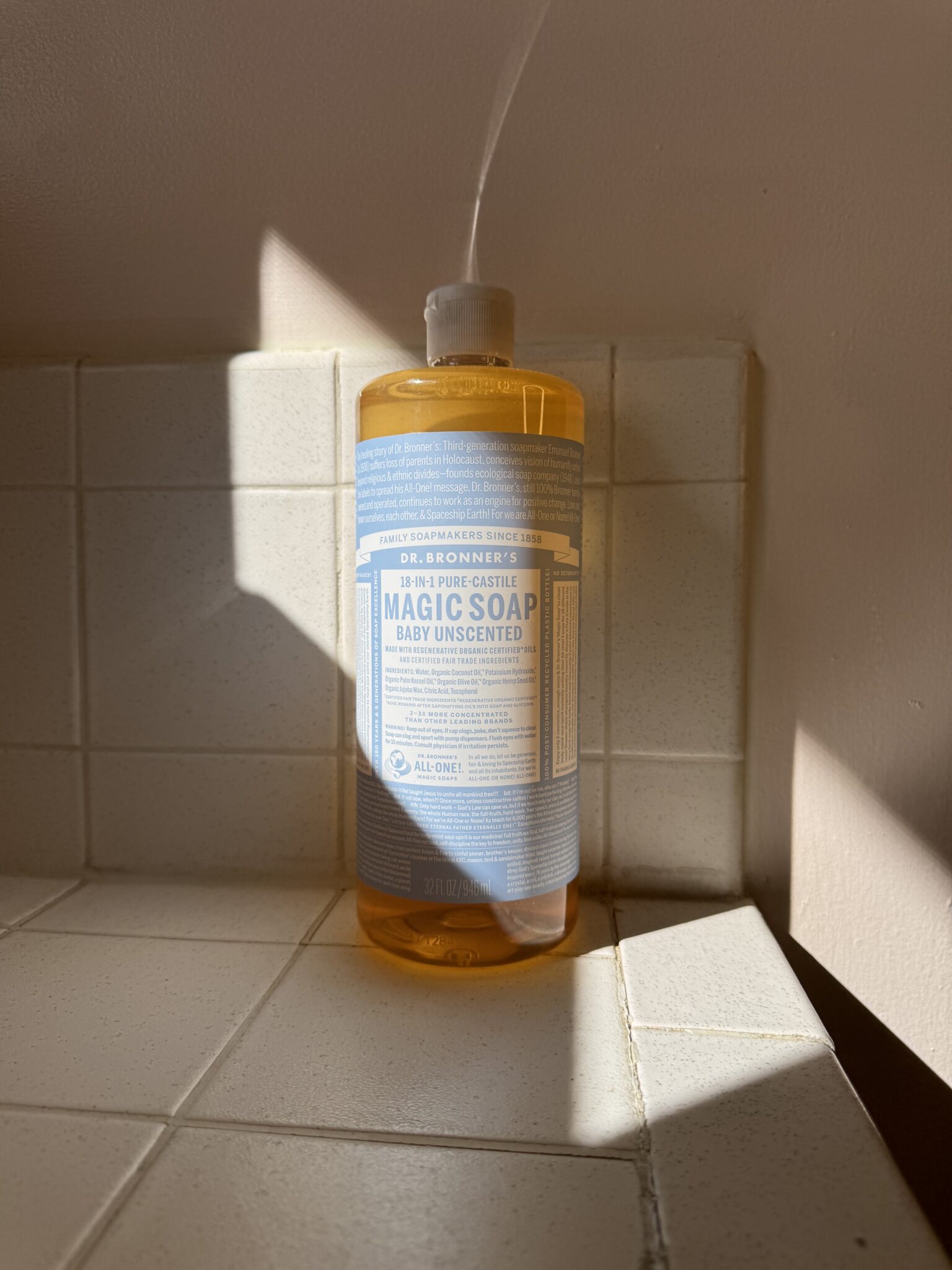 A bottle of Dr. Bronner’s 18-in-1 Pure-Castile Liquid Soap, Baby Unscented, sits on a tiled bathroom counter, illuminated by sunlight.