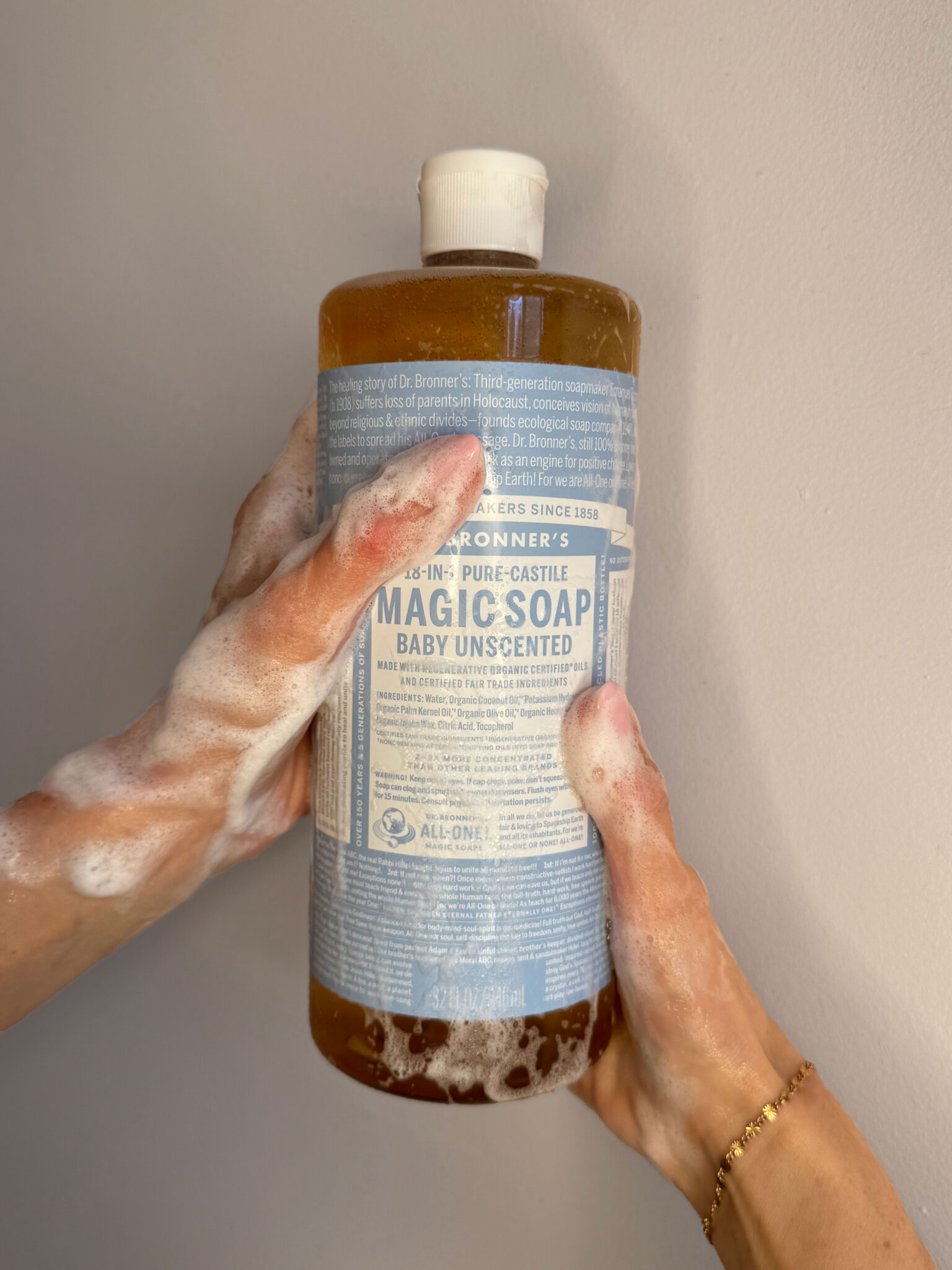 Two hands with soap lather hold a large bottle of Dr. Bronner’s Pure-Castile Baby Unscented Magic Soap against a plain light-colored background.