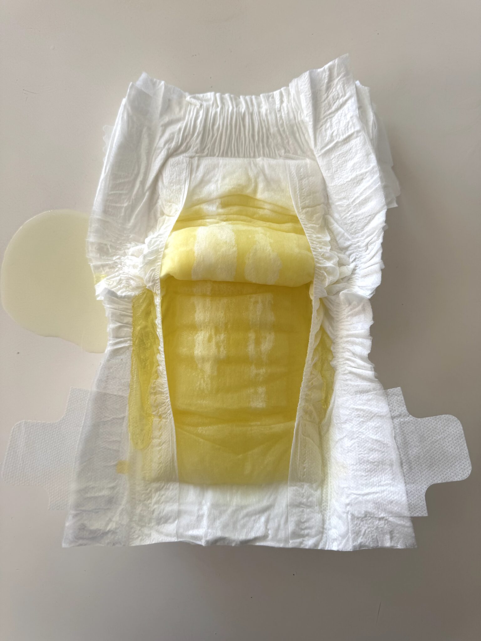 A disposable diaper with yellow liquid visibly soaked into the absorbent area and leaking slightly onto a white surface.