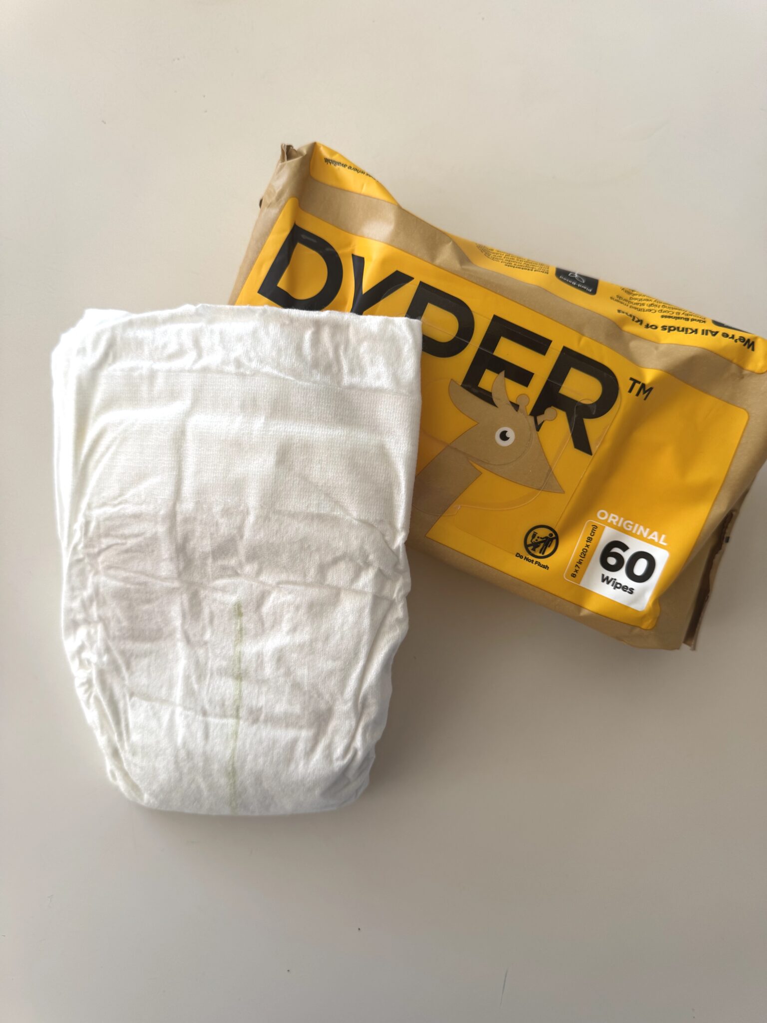 A white disposable diaper and a yellow package labeled "DYPER Original 60 Wipes" are placed on a white surface.