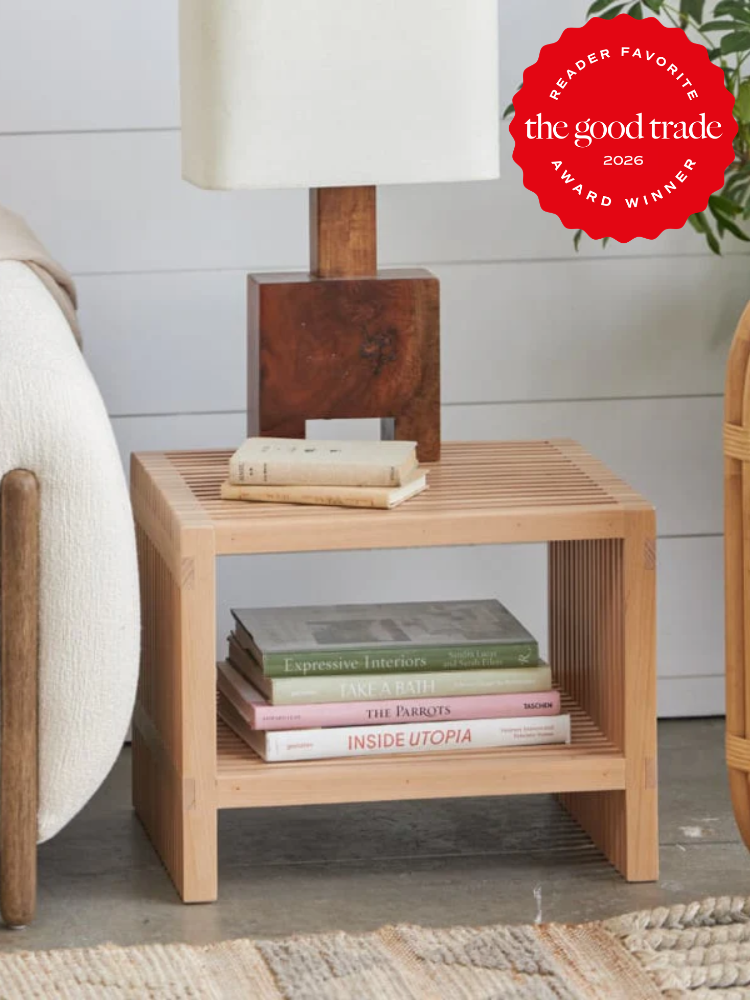 A wooden side table with a lamp on top and a stack of books on the lower shelf, next to a couch. A red "The Good Trade" award badge is in the top right corner.