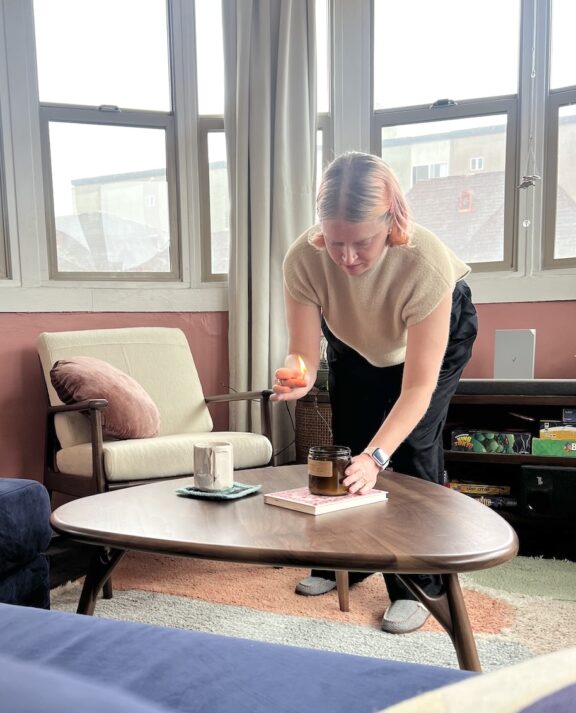 Person lighting a candle on a wooden coffee table in a living room with large windows, an armchair, and a mug nearby.