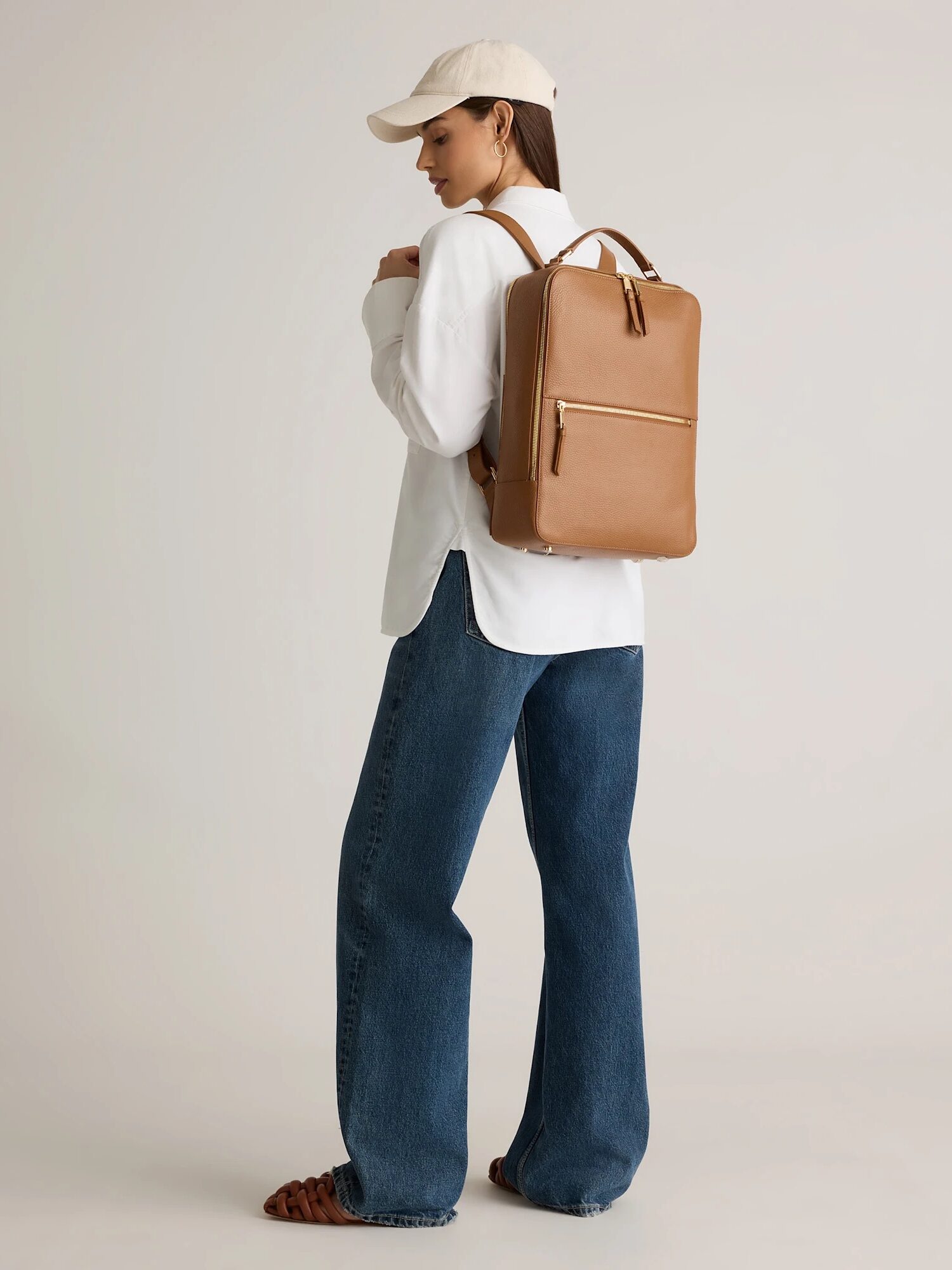 A person wearing a beige cap, white shirt, blue jeans, and brown sandals stands sideways, carrying a tan leather backpack.