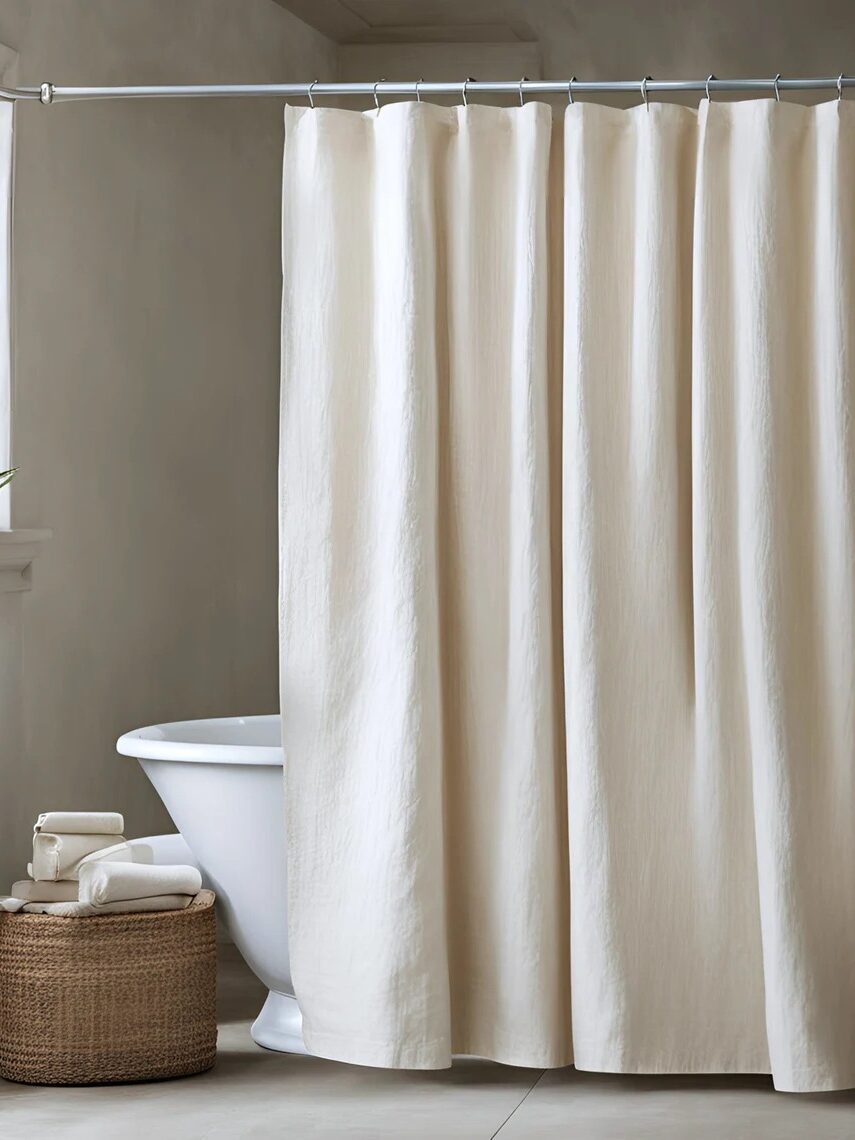 A white shower curtain hangs on a rod in front of a bathtub, with a wicker basket holding towels nearby and a small window along the wall.