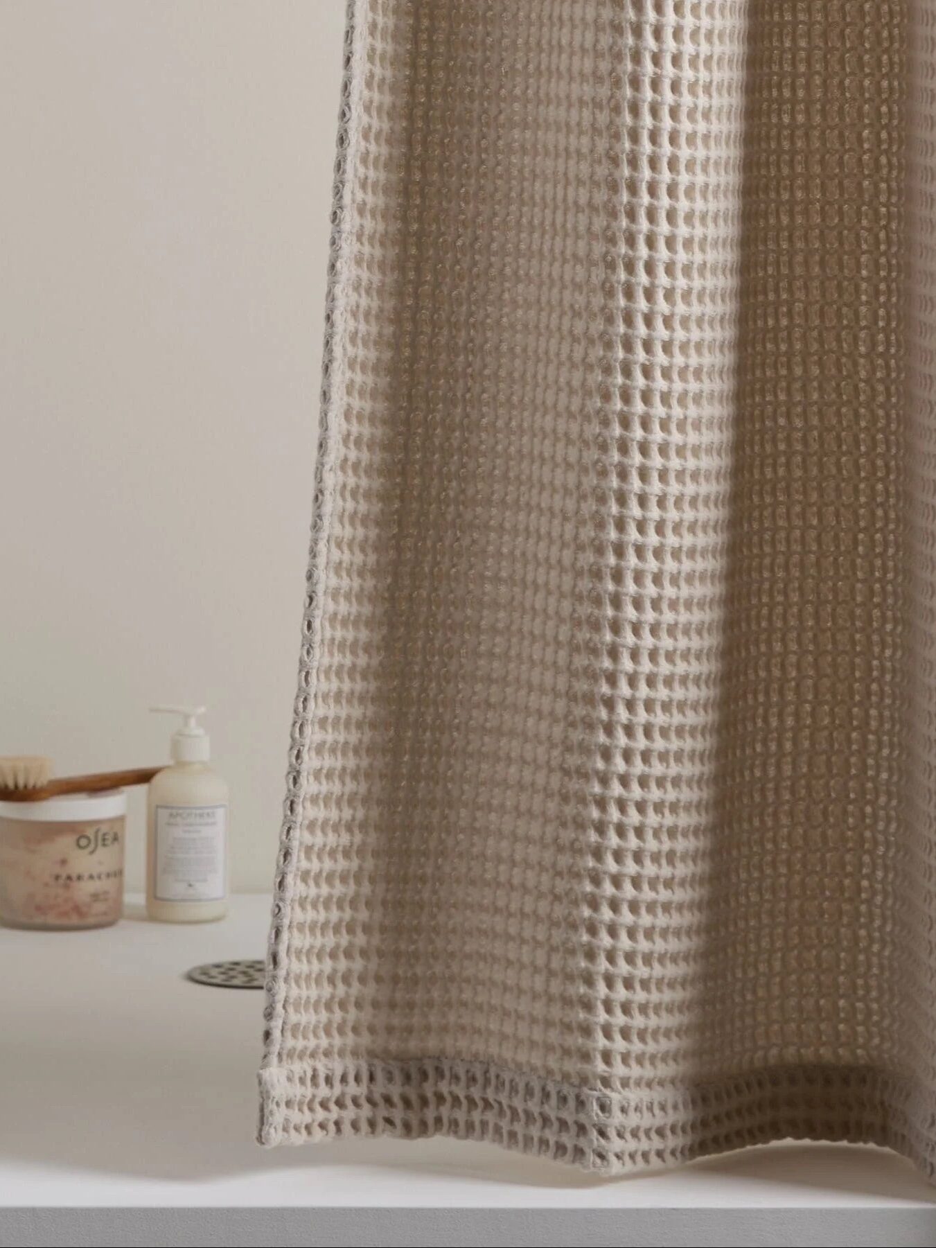 A beige waffle-textured shower curtain hangs in a bathroom; a soap dish, brush, and lotion bottle are placed on the shower ledge in the background.