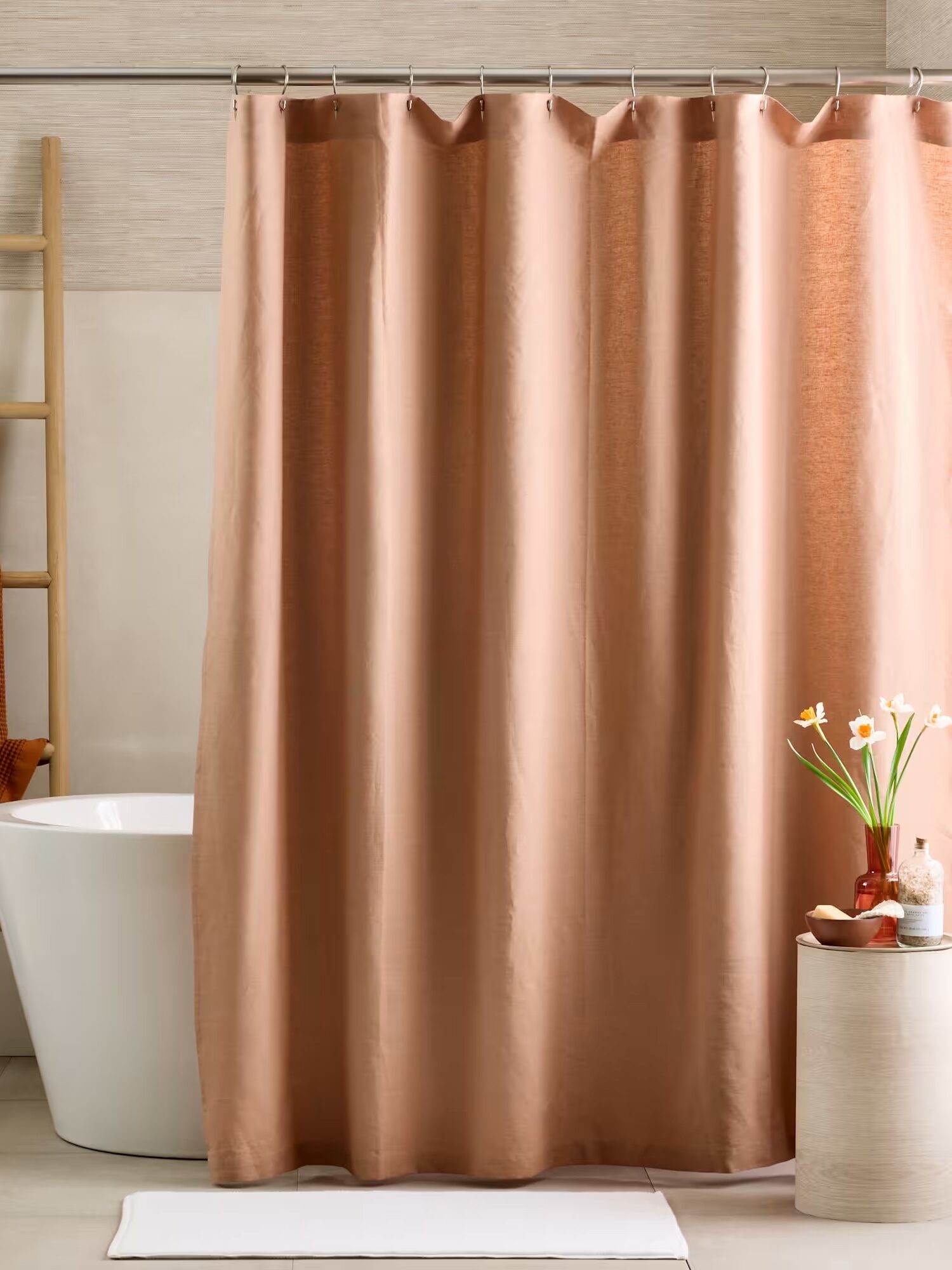 A bathroom with a freestanding tub, beige shower curtain, wooden towel ladder, orange towel, small round table with flowers, and a white bath mat on the floor.