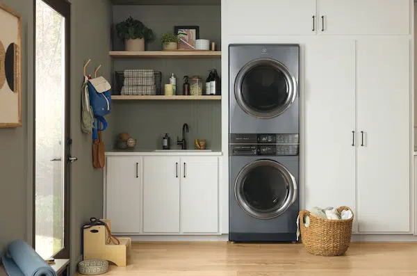 A modern laundry room with a stacked washer and dryer, white cabinets, a sink, shelves with supplies, and a basket of laundry on the floor.