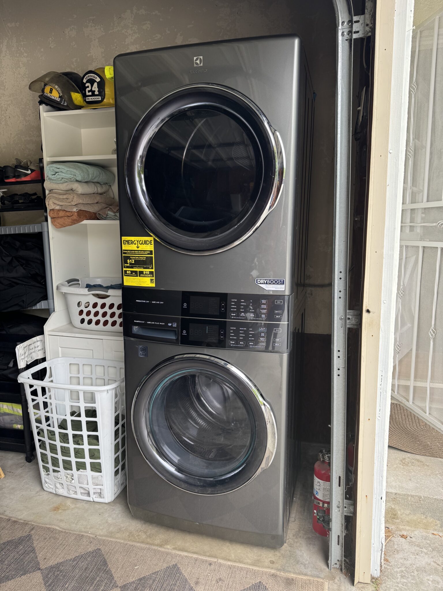 A stacked washer and dryer unit is installed next to a shelf with folded towels, laundry baskets, and a fire extinguisher in a garage or utility area.