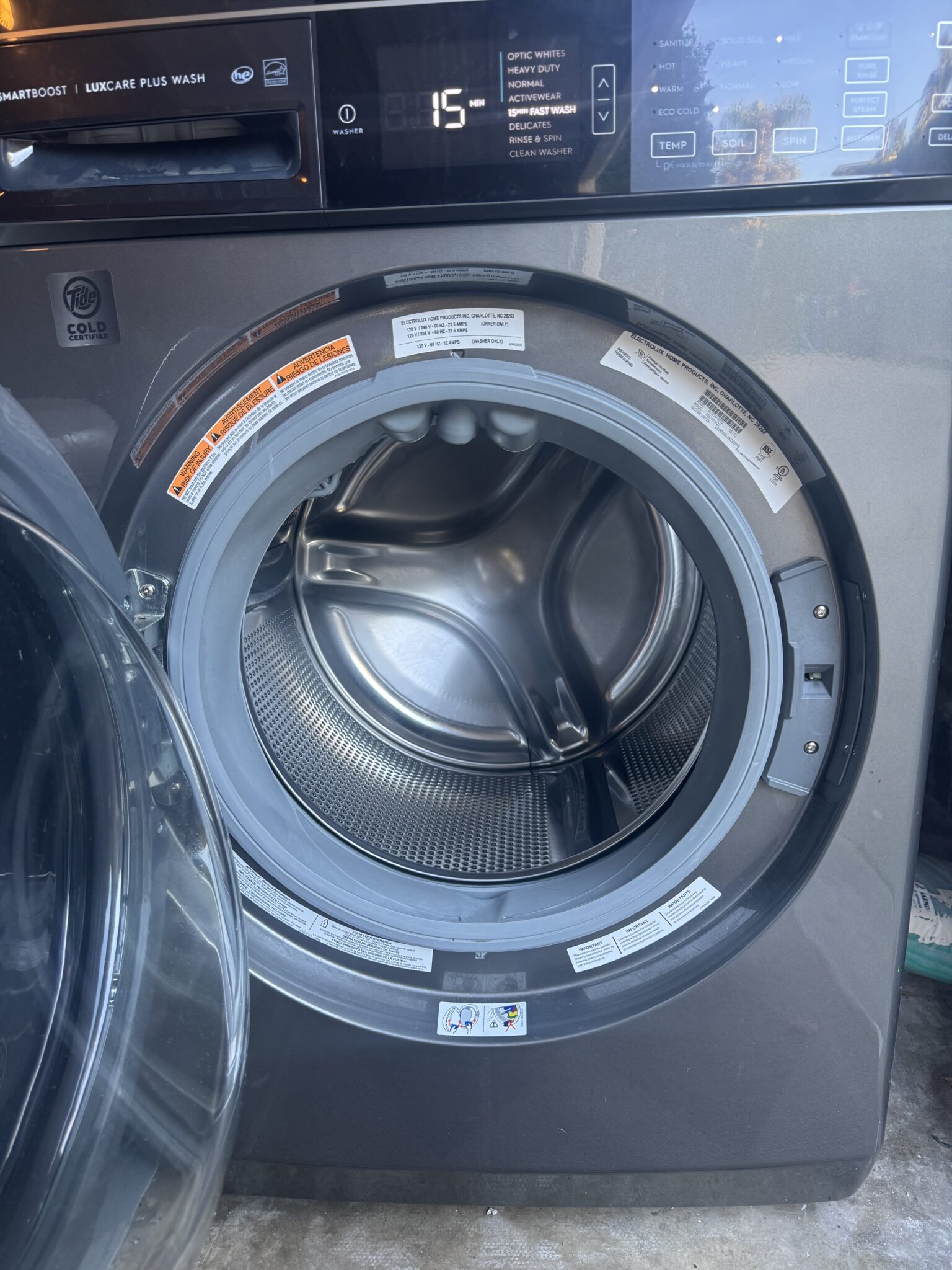 Front-loading washing machine with the door open, showing the empty drum. Control panel displays the number 15. Various warning labels are visible inside the door frame.