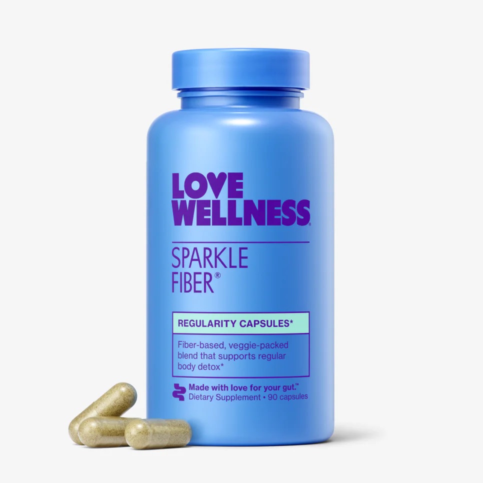 A blue bottle labeled "Love Wellness Sparkle Fiber Regularity Capsules," a popular fiber supplement for women, sits on a white background with three capsules placed beside it.