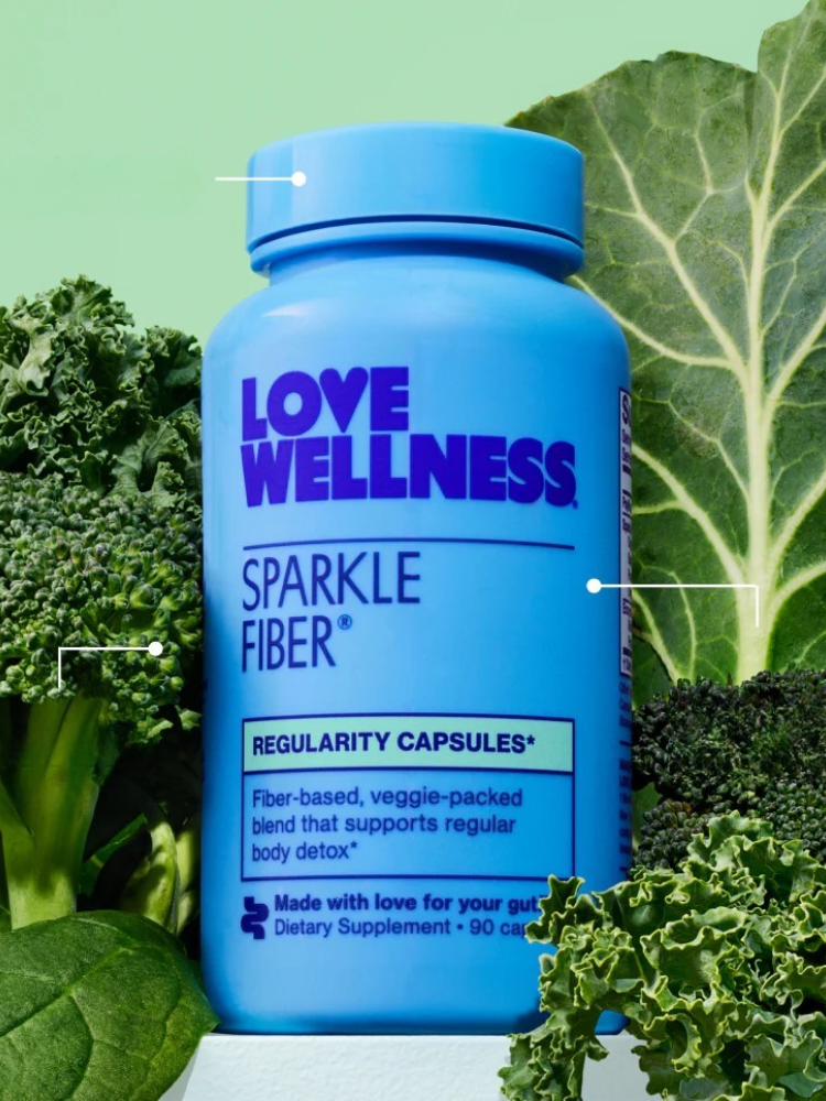 A blue bottle of Love Wellness Sparkle Fiber dietary supplement, one of the top fiber supplements for women, is displayed among green leafy vegetables like kale and collard greens.