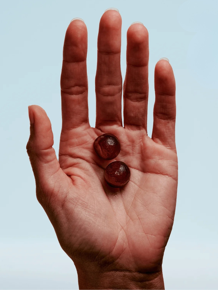 A hand is held upright with the palm facing the camera, holding two round, dark red fiber supplements for women against a light blue background.
