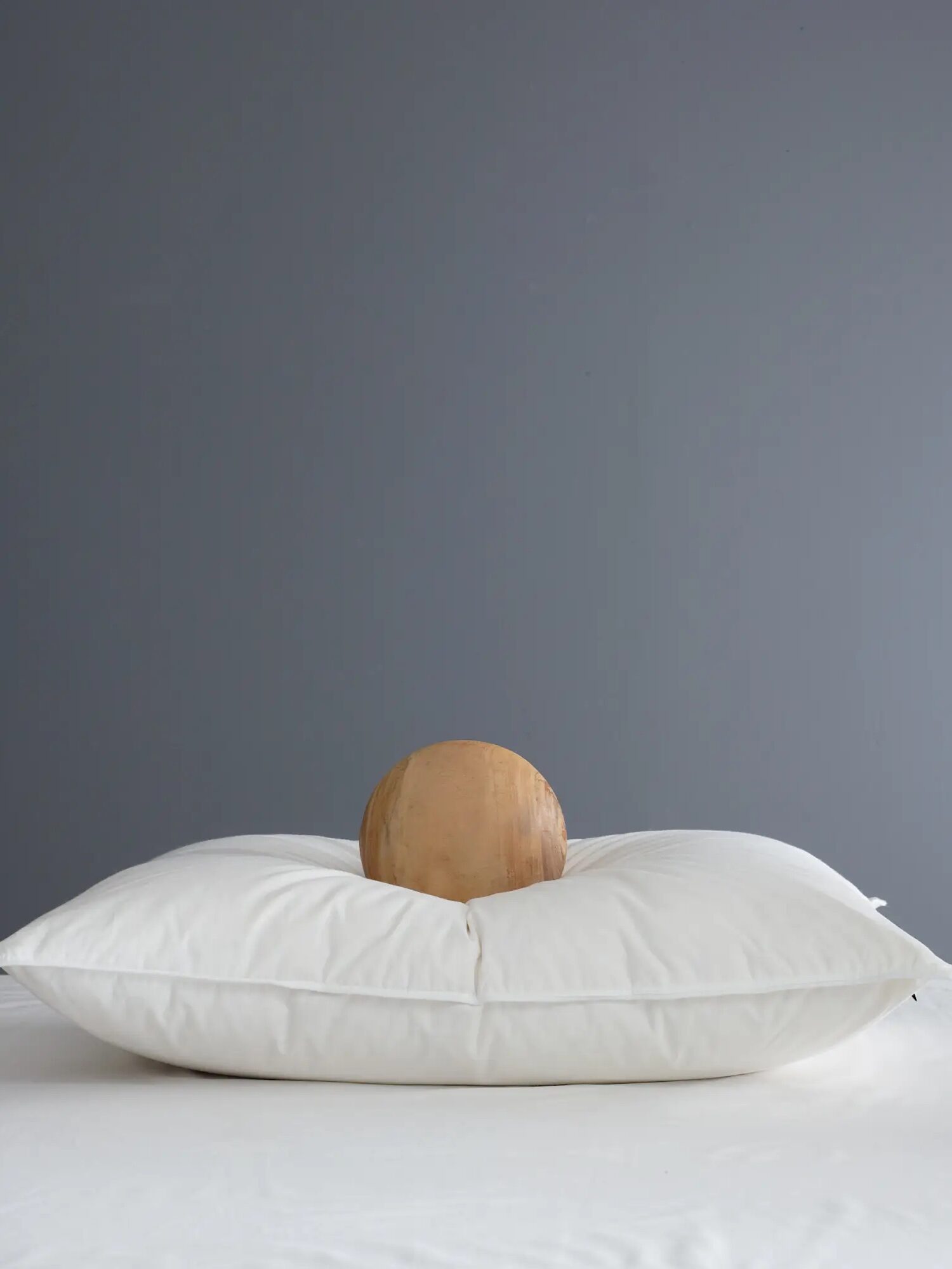 A wooden sphere rests in the center of a white pillow against a plain gray background.