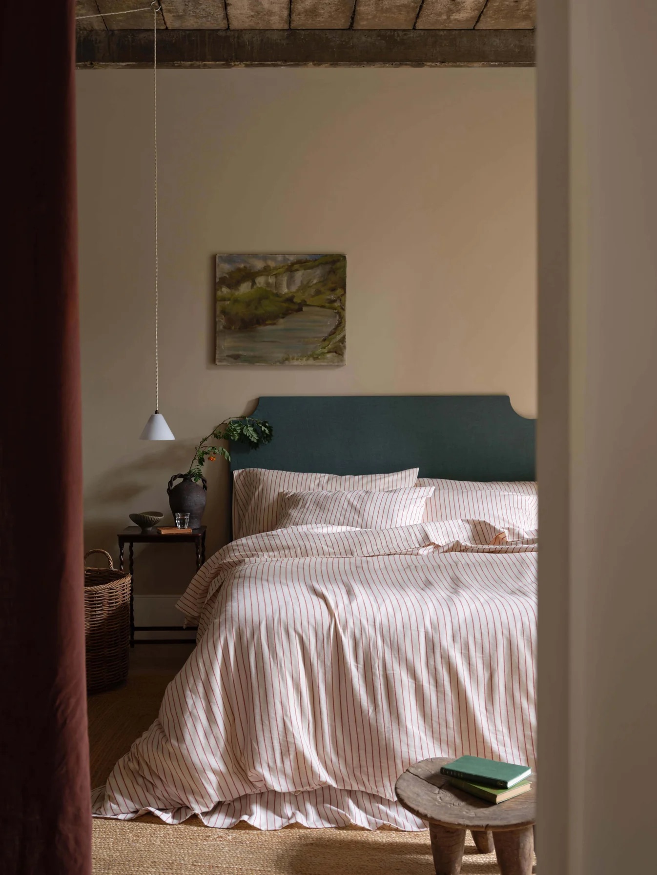 A neatly made bed with striped bedding, a green headboard, and a painting on the wall, in a softly lit bedroom with minimalist decor.