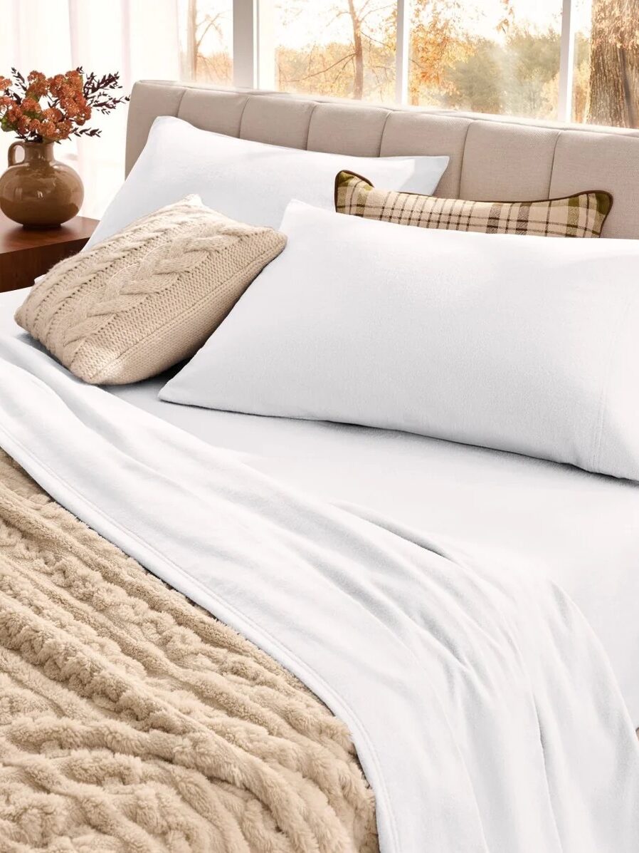 A neatly made bed with white sheets and pillows, a beige cable knit blanket, a matching pillow, and a plaid accent pillow, next to a nightstand with a vase of flowers.
