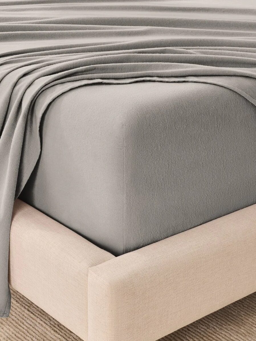 A close-up of a neatly made bed with a gray fitted sheet and matching draped blanket on a beige upholstered bed frame over a textured rug.