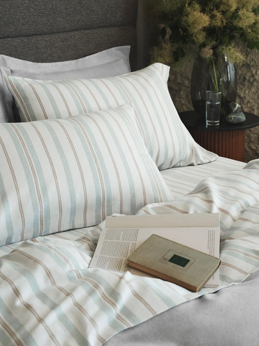 A neatly made bed with striped pillows and bedding, an open book, and a closed notebook placed on top; a glass of water and a vase with flowers are on the side table.