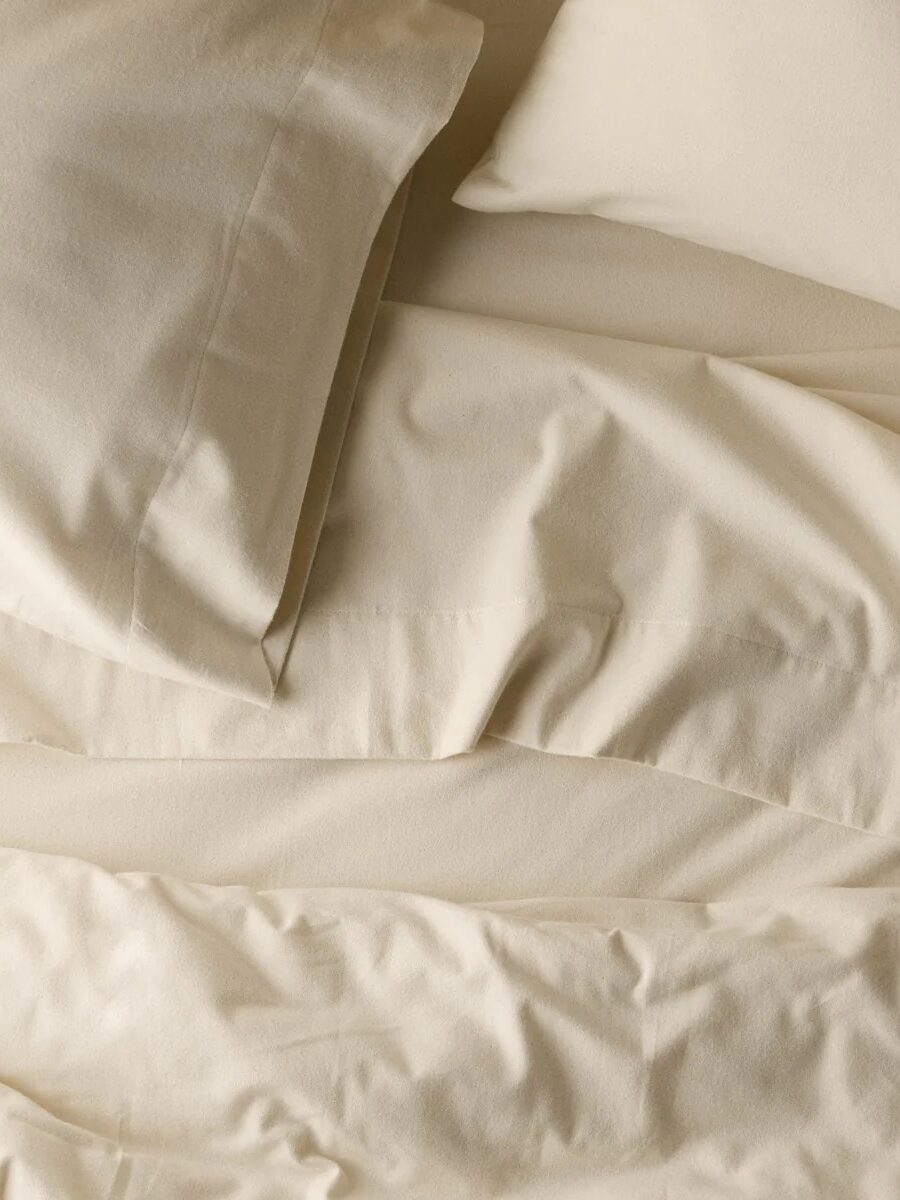 A close-up view of a bed with beige sheets and pillowcases, showing slightly wrinkled fabric and an unmade appearance.