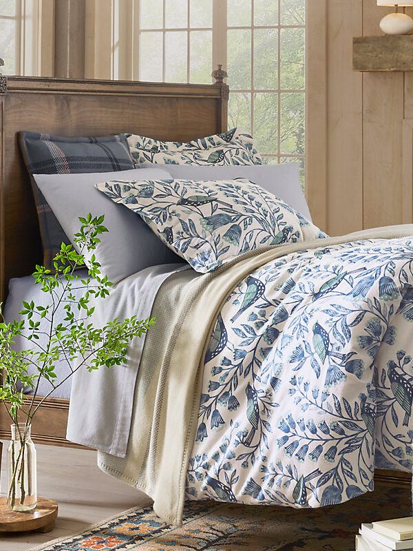 A neatly made bed with blue and white floral bedding, gray sheets, and multiple pillows, placed in a cozy wooden room with large windows and a potted plant nearby.