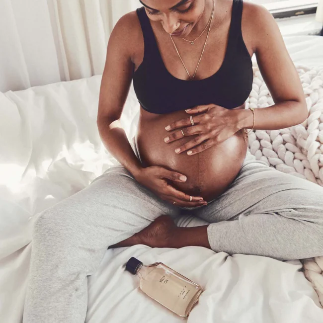 Pregnant person sitting on a bed in comfortable clothes, holding their belly with both hands; a bottle of body oil lies nearby.