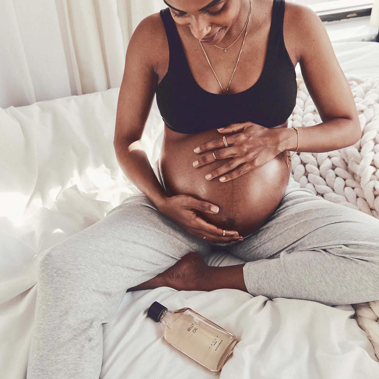 Pregnant person sitting on a bed in comfortable clothes, holding their belly with both hands; a bottle of body oil lies nearby.