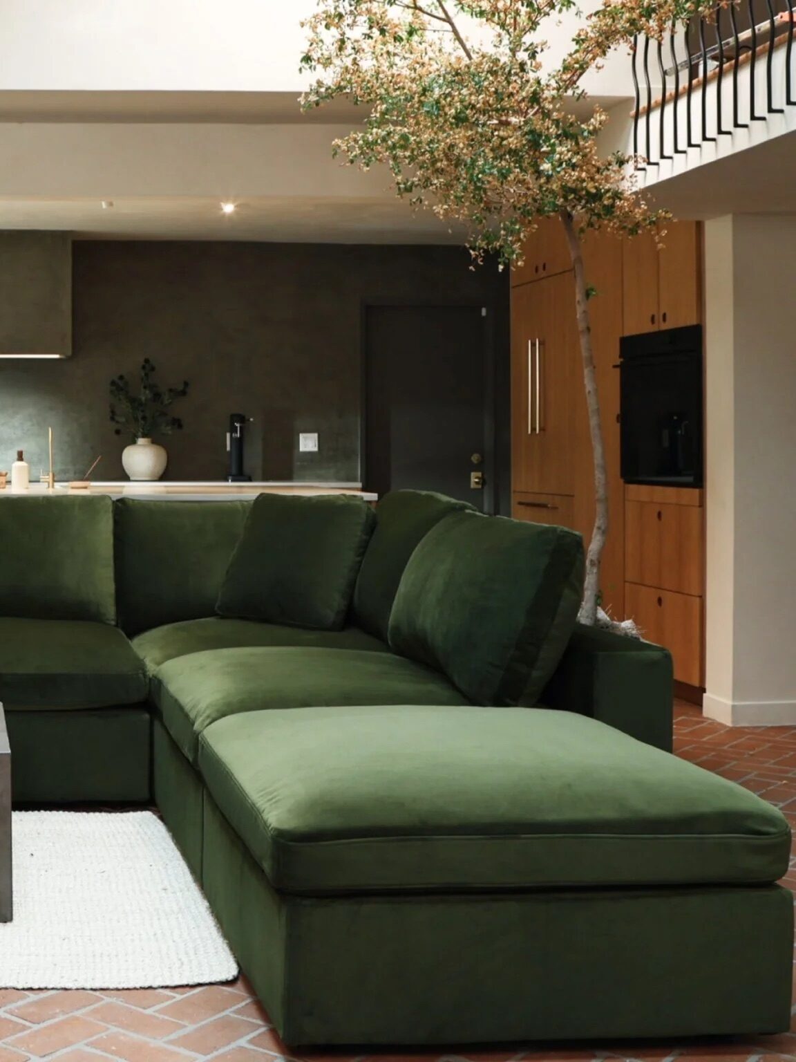 A large green sectional sofa sits on a white rug in front of a low gray coffee table in a modern, open-concept living room and kitchen.