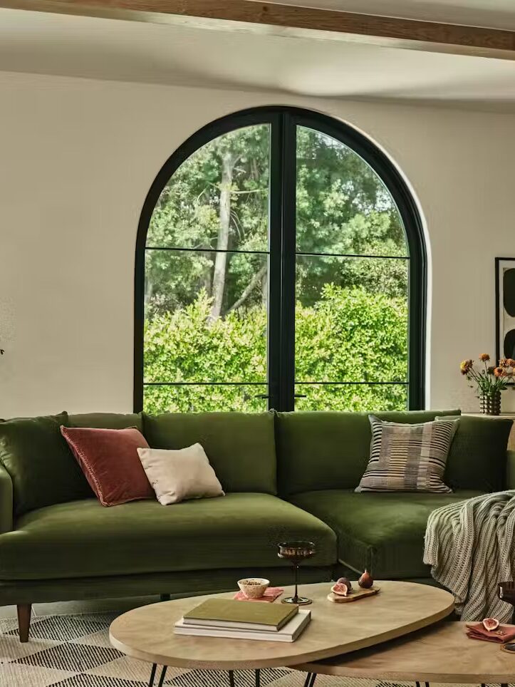 A modern living room with a green sectional sofa, a large arched window, a potted plant, wooden coffee tables, and a sideboard with decor against a white wall.