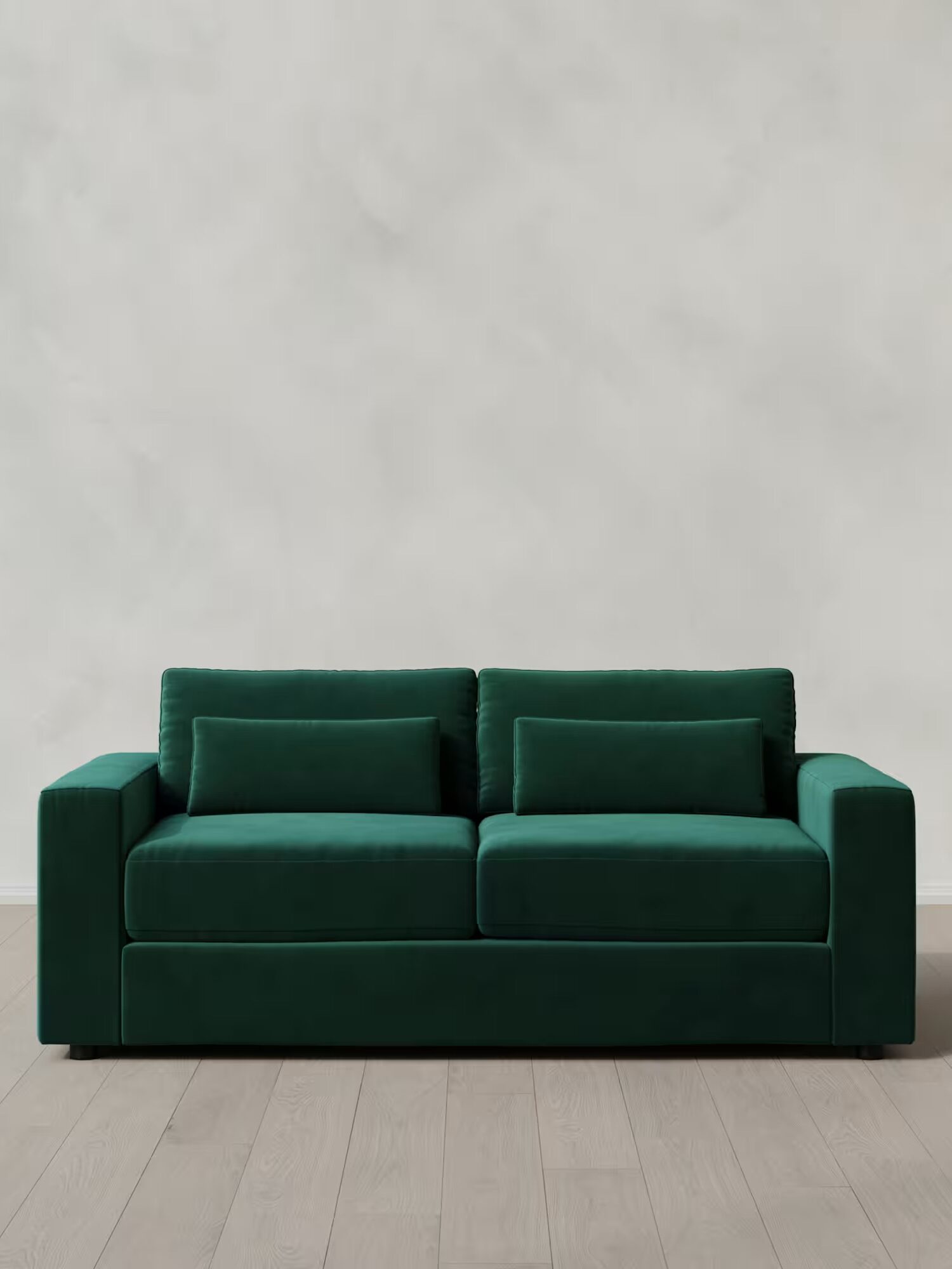 A green velvet sofa with two matching cushions sits against a plain, light-colored wall on a light wooden floor.