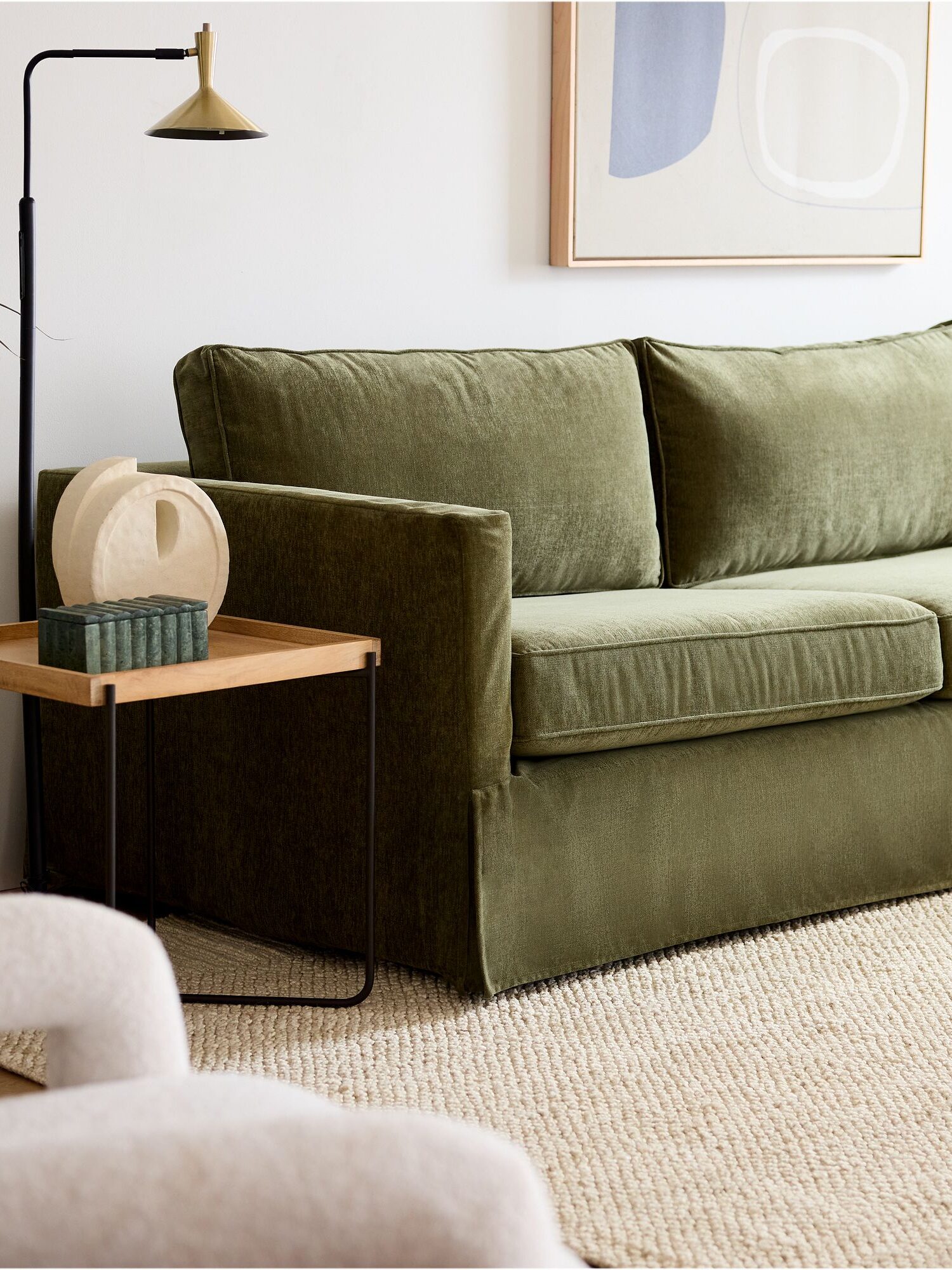 A green sofa sits next to a wooden side table with decorative items, a floor lamp, and abstract art on a white wall, with a beige textured rug on the floor.