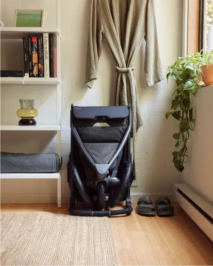A folded black stroller is leaned against a wall below a hanging robe, next to shelves and a window with a potted plant.