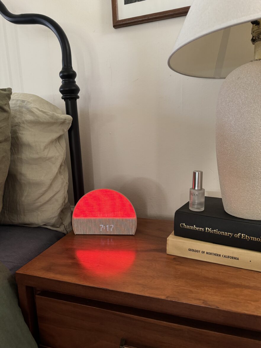 A bedside table with a digital clock glowing red, a small bottle, two stacked books, part of a lamp, and the edge of a bed with pillows.
