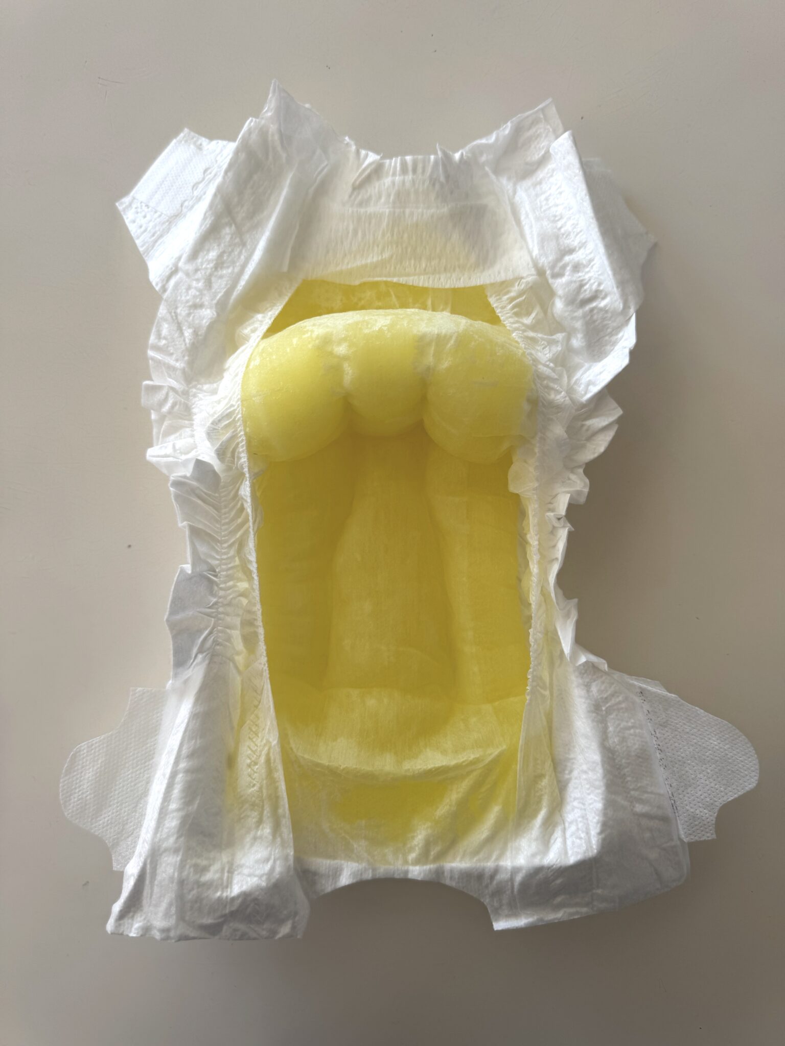 An open disposable diaper with a visible yellow liquid absorbed in the center, displayed on a neutral background.
