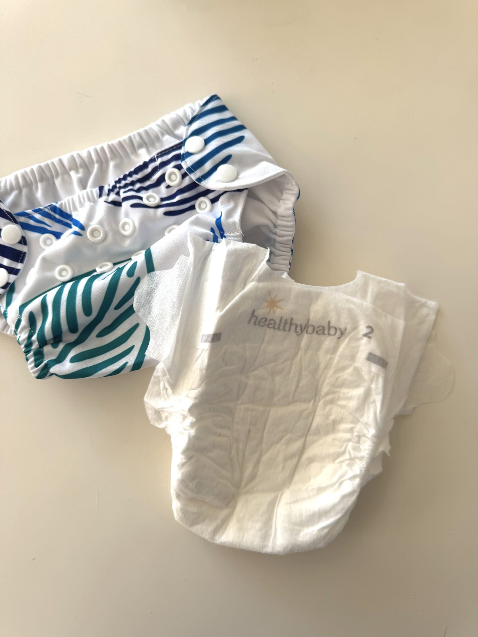 A reusable cloth diaper with blue and green stripes sits next to a disposable HealthyBaby diaper, size 2, on a white surface.