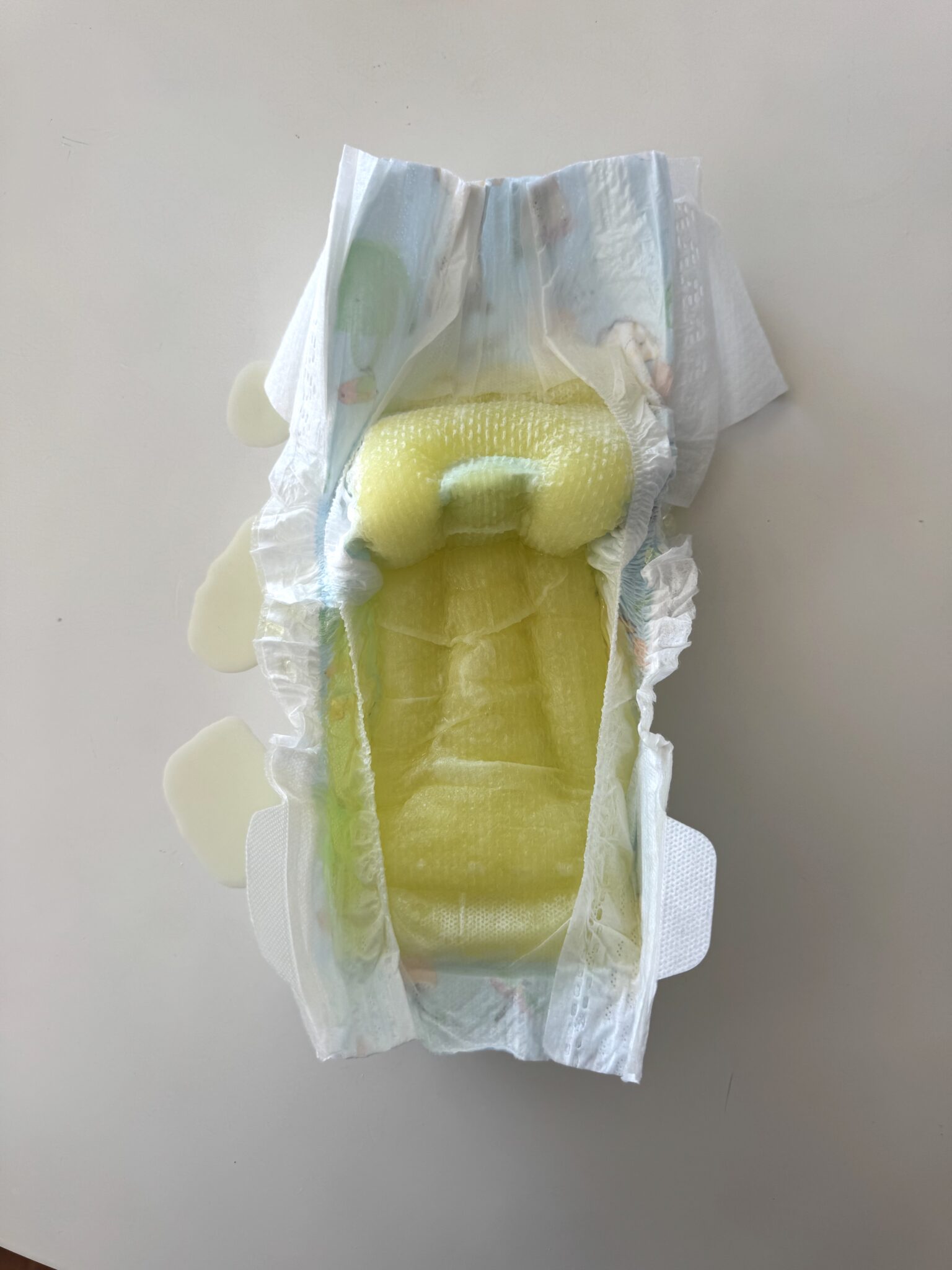 An opened disposable diaper with yellow liquid absorbed inside, showing leakage on the surrounding white surface.