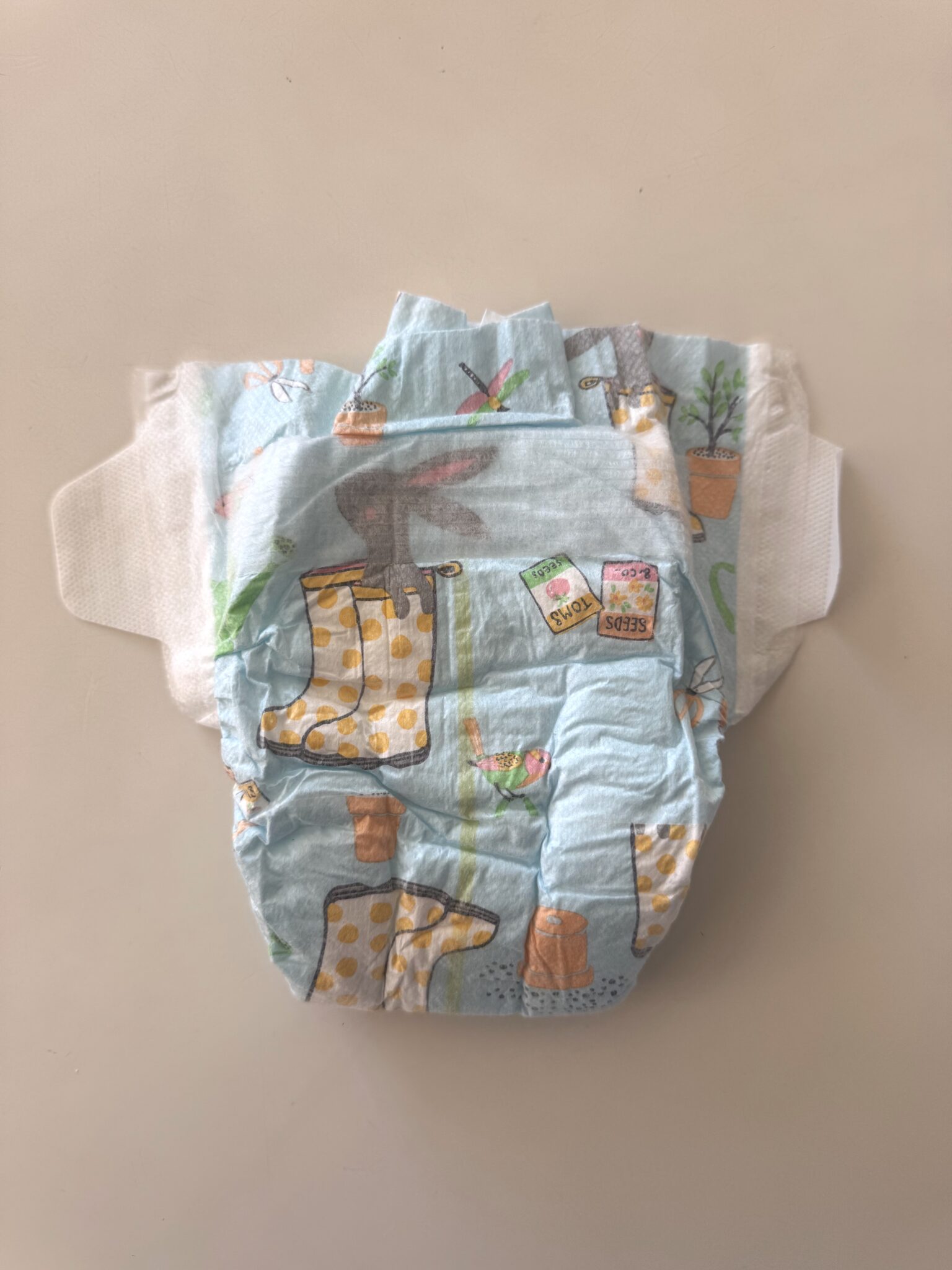 A disposable baby diaper with colorful printed designs, including yellow rain boots, plants in pots, and seed packets, lying on a plain surface.