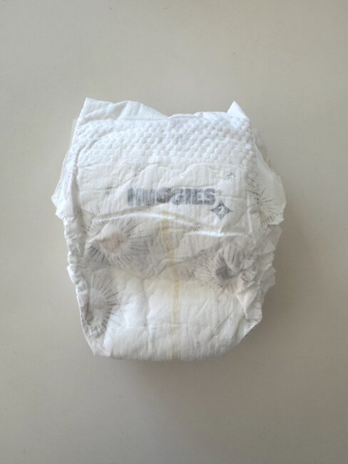 A used disposable Huggies diaper with a visible yellow stain is placed on a plain, light-colored surface.
