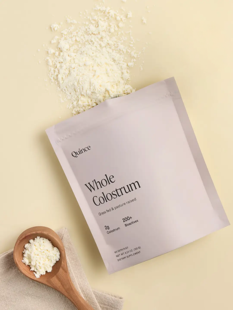 A packet labeled "Whole Colostrum" lies on a beige surface with colostrum powder scattered nearby and a wooden spoon holding more powder rests on a cloth.