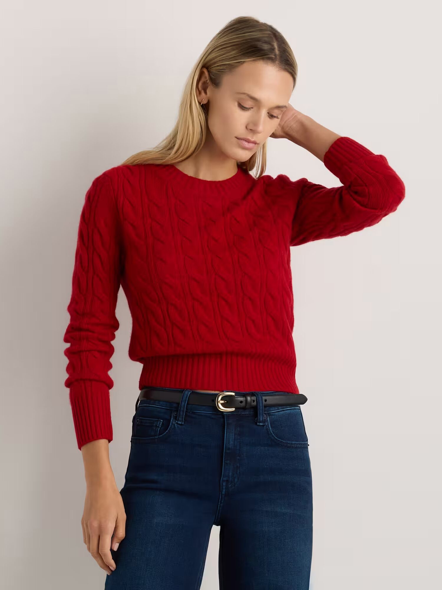 A woman wearing a red cable-knit sweater and dark blue jeans stands with one hand touching the side of her head against a plain light background.