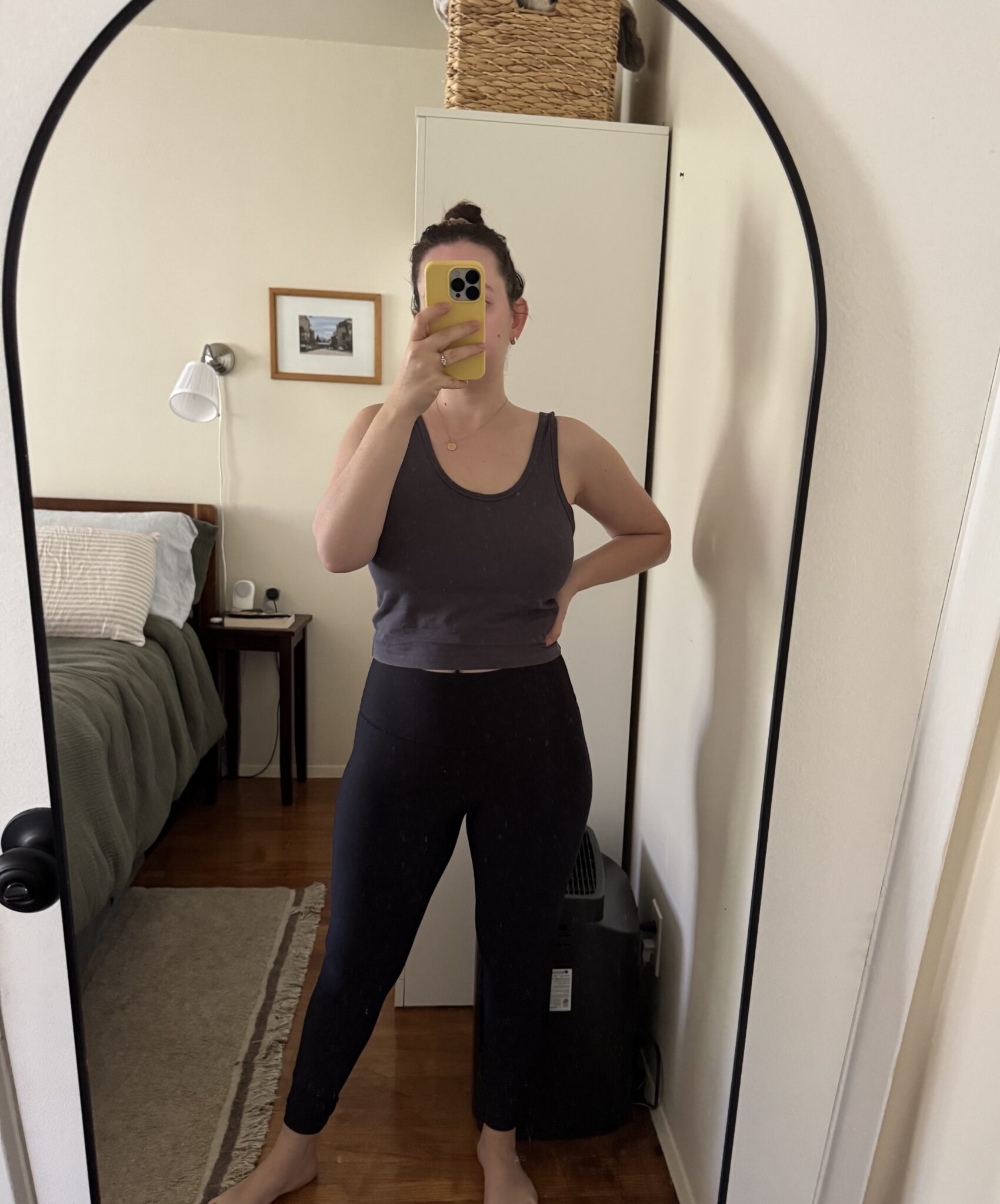 A person in a dark tank top and leggings takes a mirror selfie in a bedroom with a made bed, bedside table, and a basket on top of a cabinet.