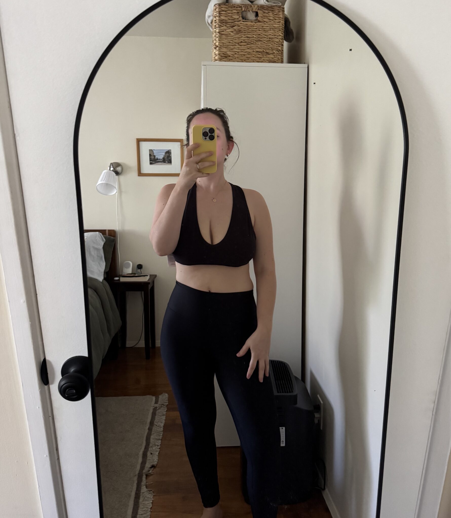 A person wearing a black sports bra and black leggings takes a mirror selfie in a bedroom with a bed, nightstand, and wall art visible in the background.