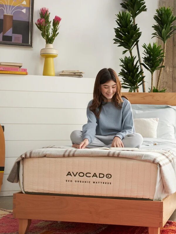 A woman sits cross-legged on a bed with an Avocado Eco Organic Mattress in a bright bedroom with large plants, a skateboard, and modern decor.