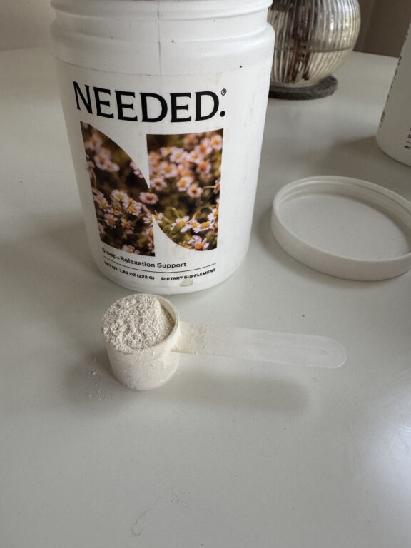 A white container labeled "NEEDED." sits open on a table with a white plastic scoop filled with powder resting in front of it.