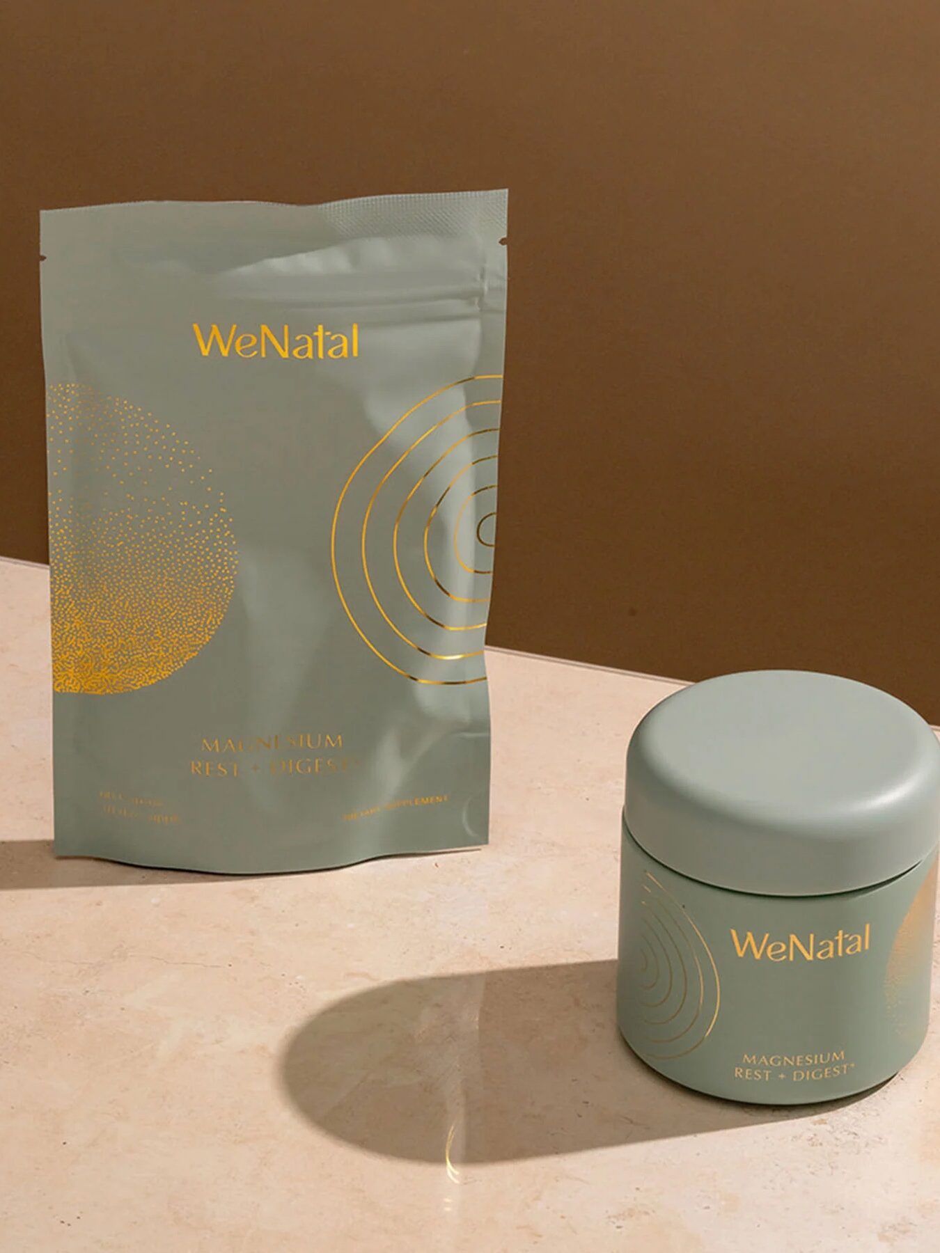 A light green pouch and matching jar labeled "WeNatal Magnesium Rest & Digest" on a beige surface with a brown background.