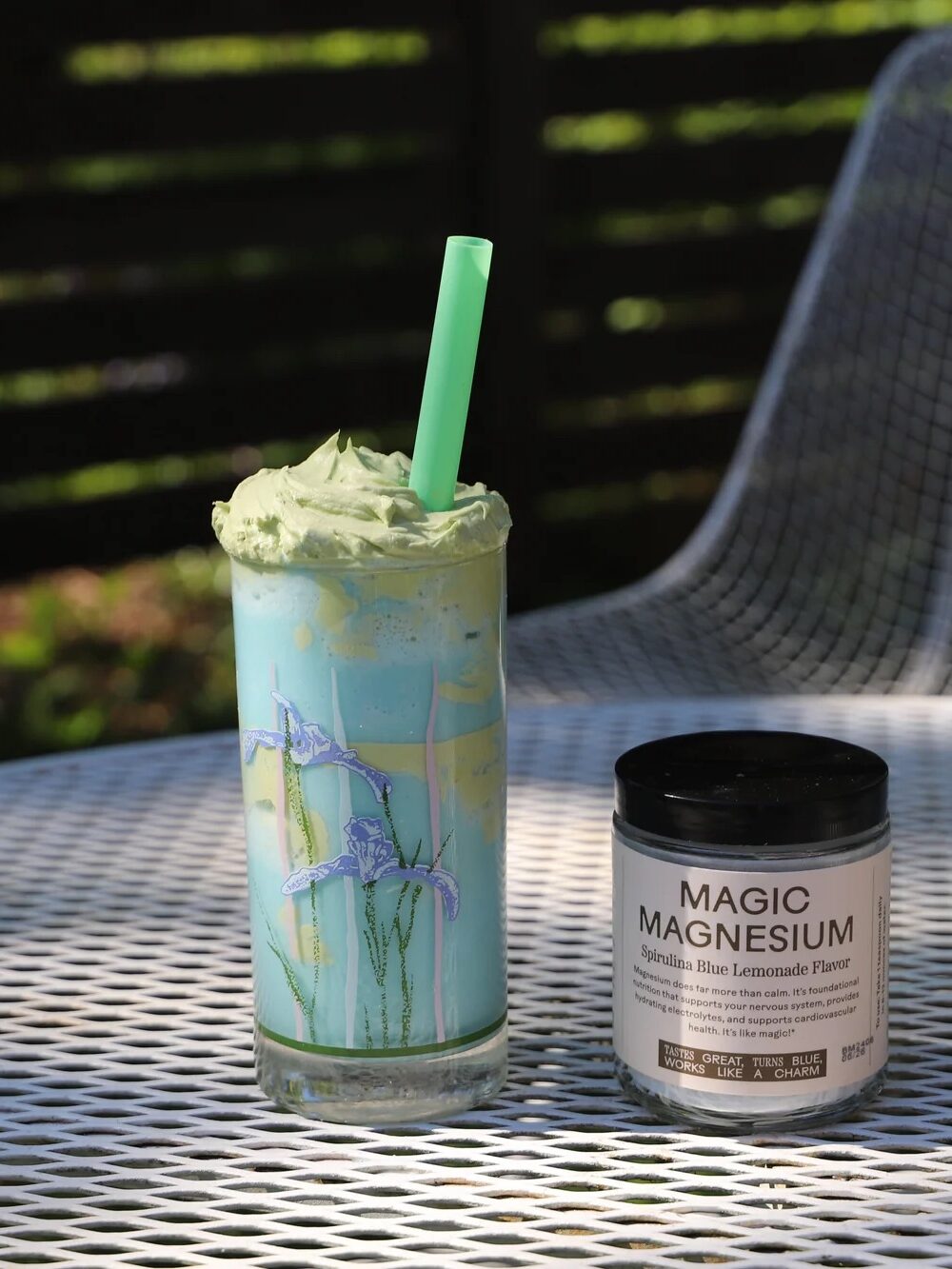 A glass of green smoothie with a straw sits on a white outdoor table next to a jar labeled "Magic Magnesium." Two wire mesh chairs are in the background.