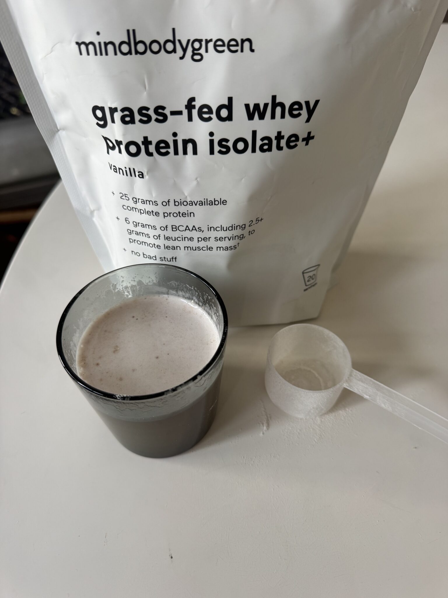 A glass of mixed protein shake sits next to an empty scoop and a bag labeled "mindbodygreen grass-fed whey protein isolate+ vanilla.