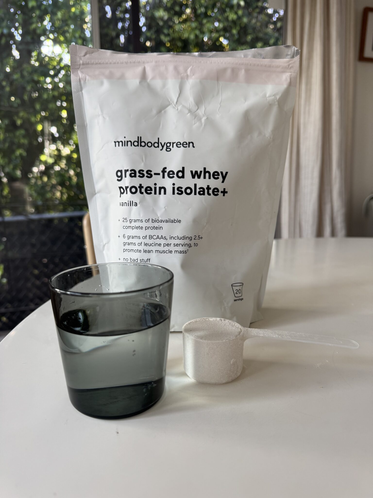 A glass of water, a scoop of protein powder, and a bag labeled “mindbodygreen grass-fed whey protein isolate+ vanilla” are on a white table.