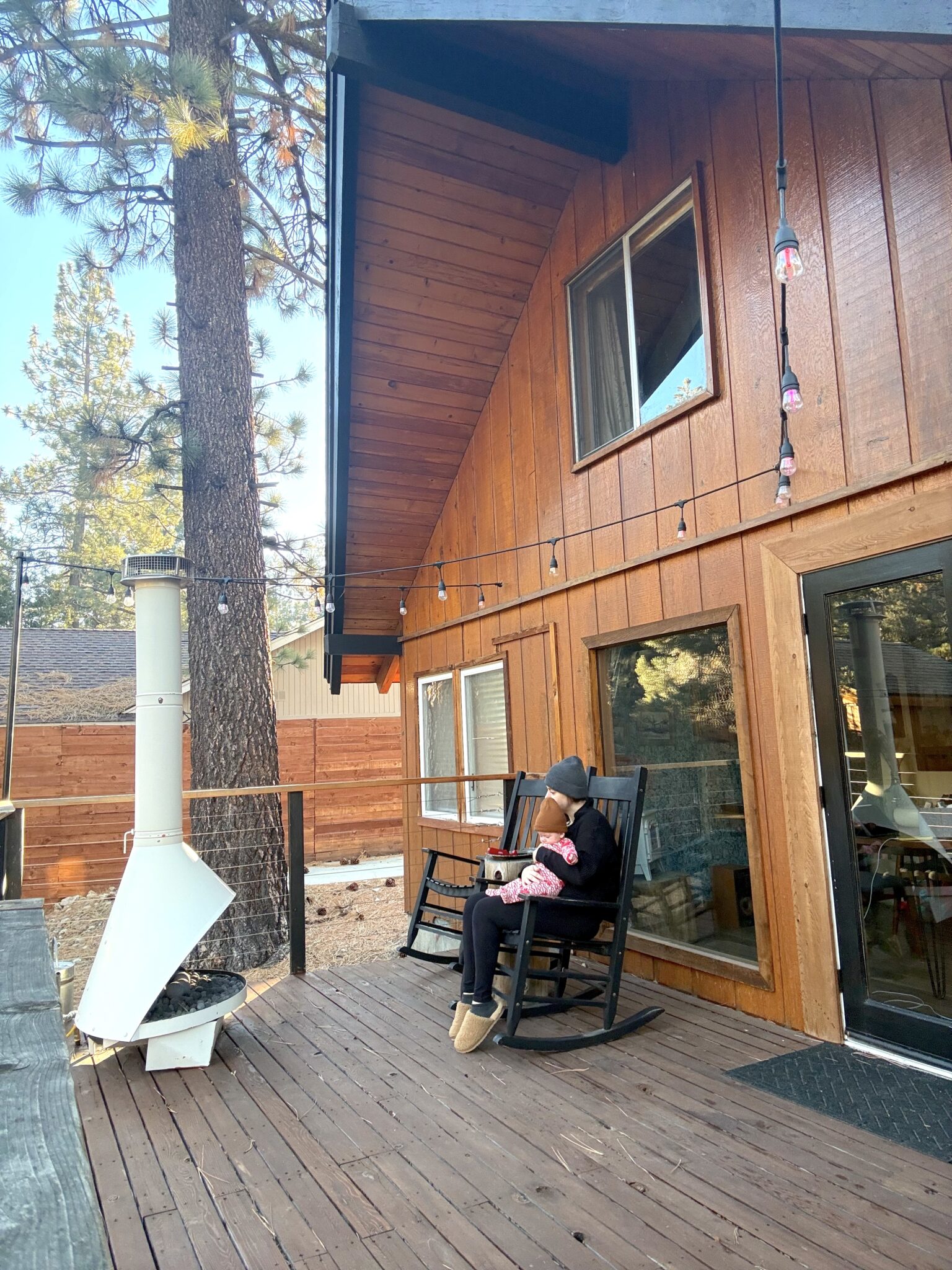 A person sits in a rocking chair holding a baby on the wooden deck of a cabin with large windows, surrounded by trees and outdoor string lights.