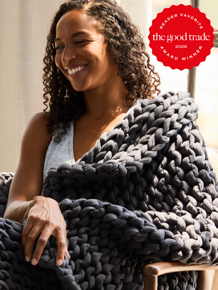 A woman sits smiling, wrapped in a chunky dark gray knit blanket. A red “The Good Trade Reader Favorite Award Winner 2026” badge appears in the top right corner.